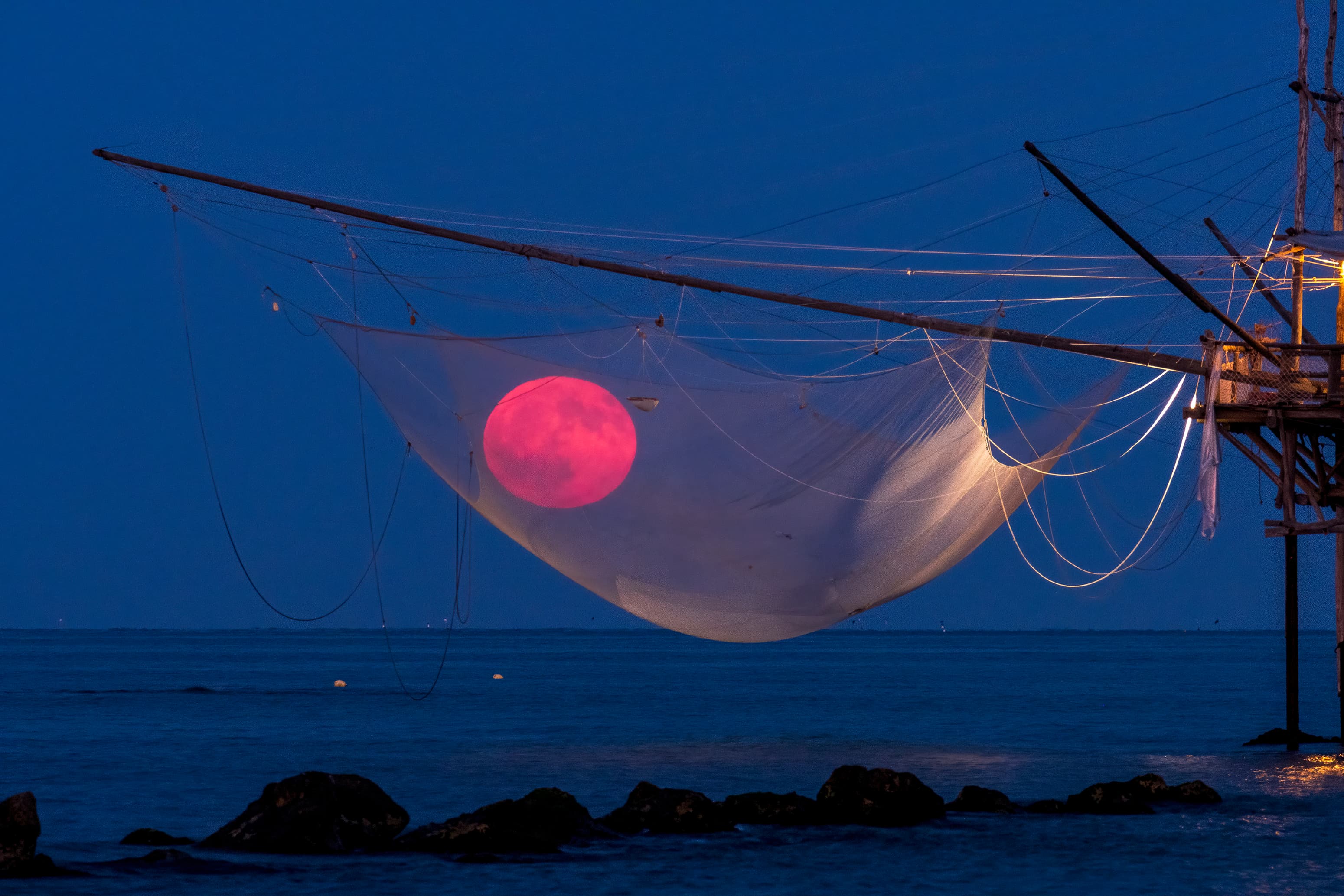 قمر محمرّ يعلو الأفق بقليل يظهر من خلال شبكة صيد تتدلّى من منصّة تعلو شاطئاً ترتصف فيه بعض الصخور
