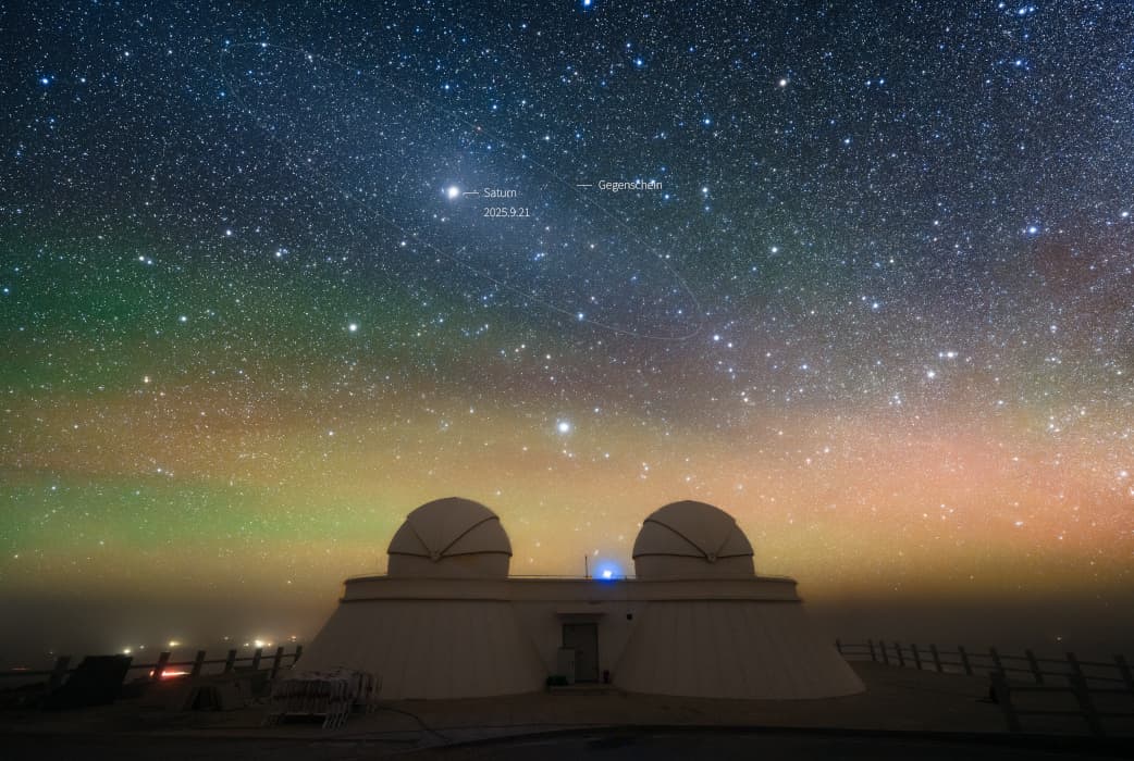 سماء ليلٍ بألوانٍ متماوجة ترصّعها النجوم ويظهر فيها جرمٌ ساطعٌ هو زُحل يتوسّط منطقة ضياء نسبي وتحت ذلك كلّه مُنشأة رصد بقبّتين