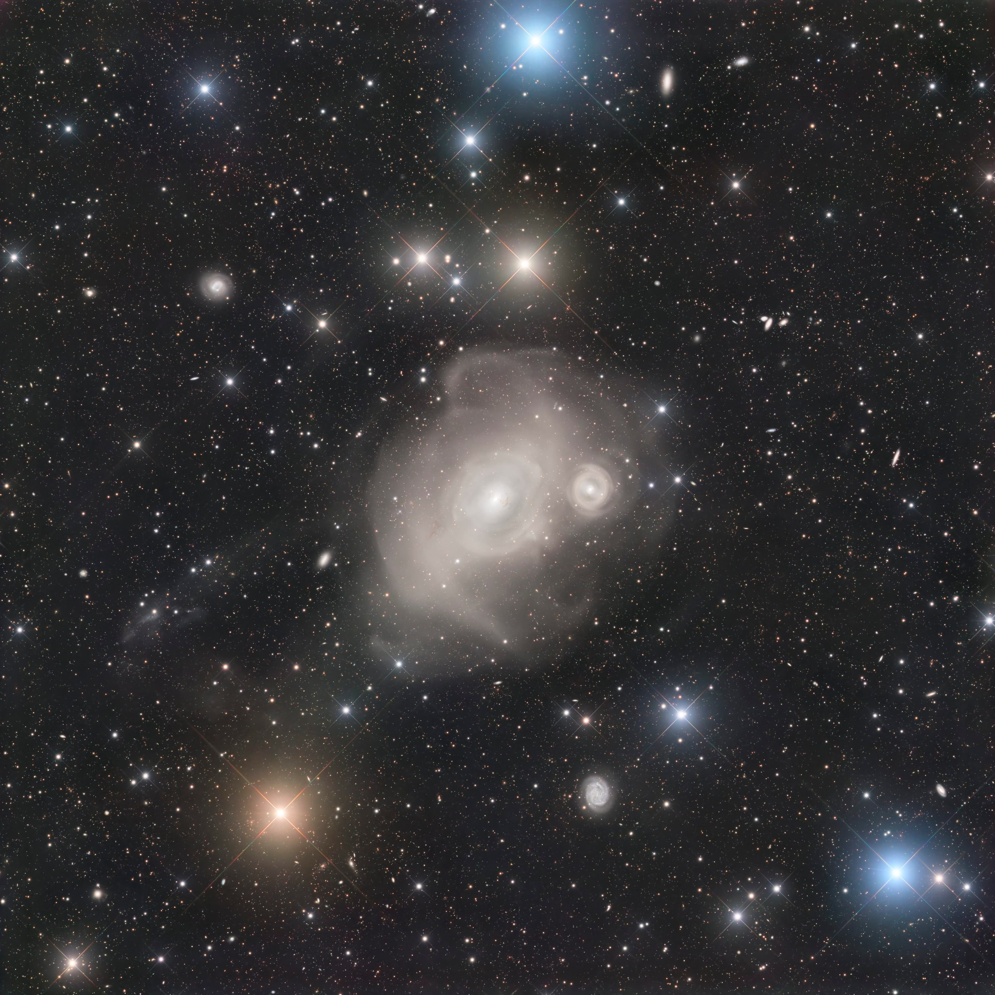 مجرّة اهليلجيّة تظهر كطبقات غباريّة ذات مركز ساطع وبجانبها مجرّة مشابهة أصغر فيما تتناثر عبر الصورة مجرّات الخلفيّة ونجوم المقدّمة