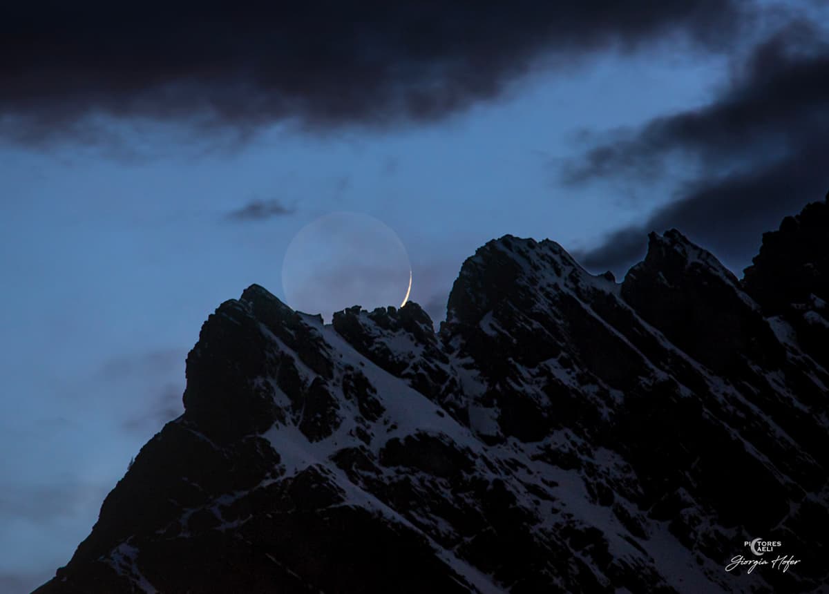 حرفٌ جبليٌّ وعرٌ بثلوج يظهر خلفه قمرٌ بهلالٍ ساطع وقرصٍ خافت وبينهما سحب 