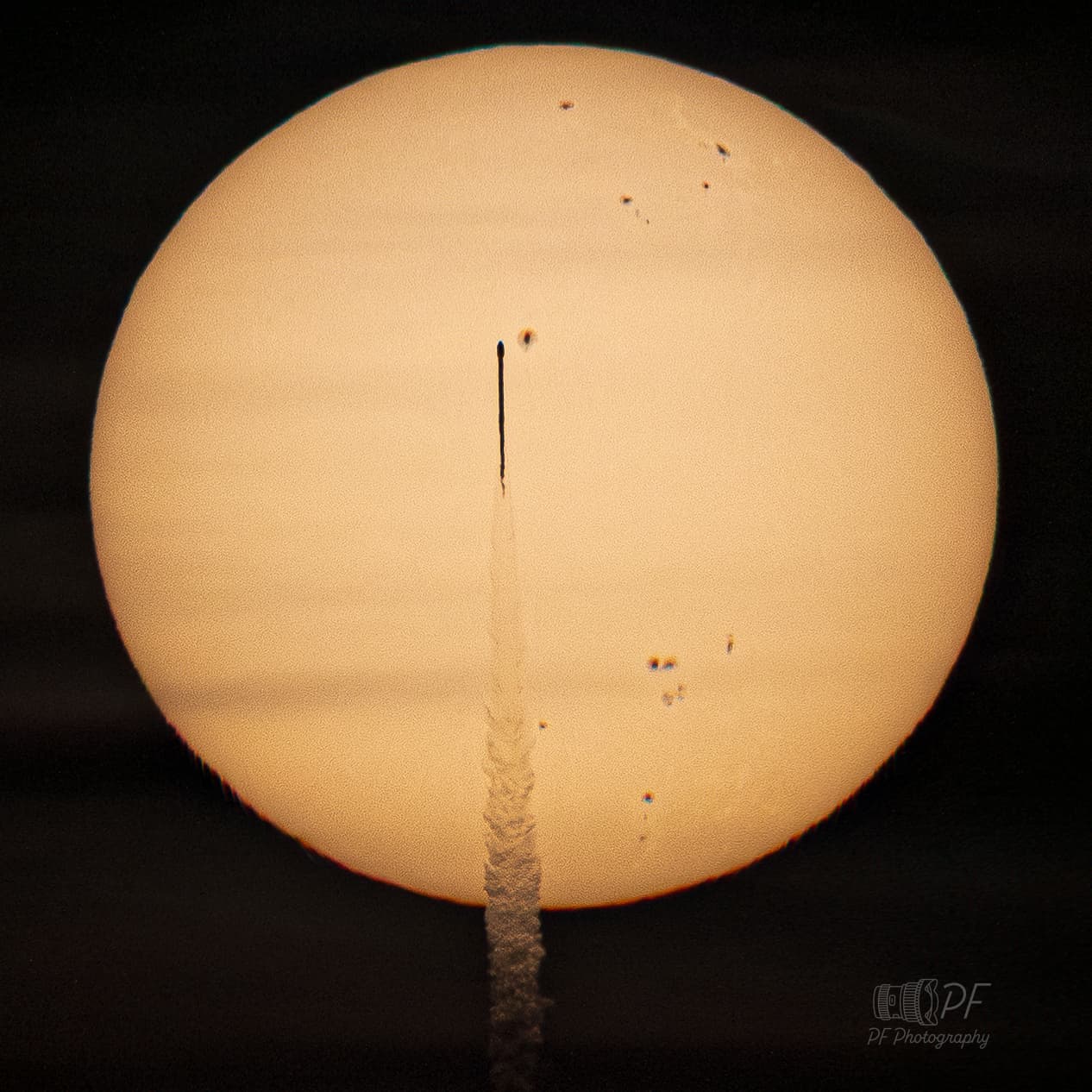 قرص الشمس وعليه بقع شمسيّة عديدة ويعبره صاروخٌ يبدو كعود ثقاب ظليل وخلفه ذيلٌ من الغازات المخلخلة للرؤية