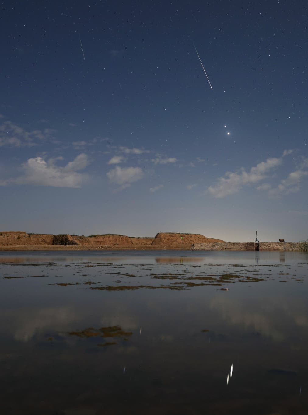 سماء تتناثر فيها النجوم وفيها جرمان ساطعان هما المشتري والزهرة ويخطّها شهابان وتعبرها سحب خفيفة وفي المقدّمة مسطّح مائي تنعكس عليه