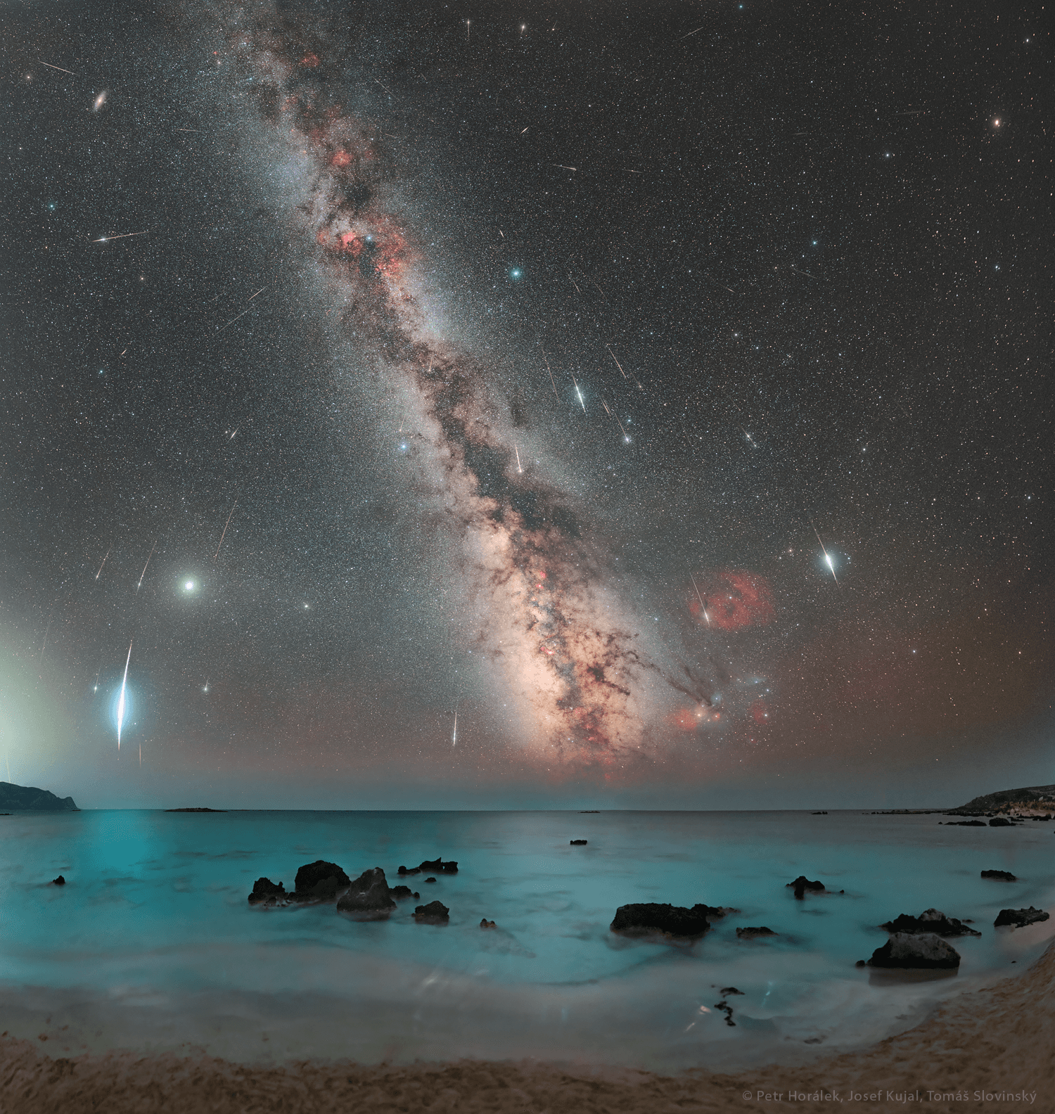 سماء مرصّعة بالنجوم يعبرها نطاق درب التبّانة وتخطّها شهب متفاوتة فيما يظهر في المقدّمة شاطئ وبحر أزرق فيه بعض الصخور