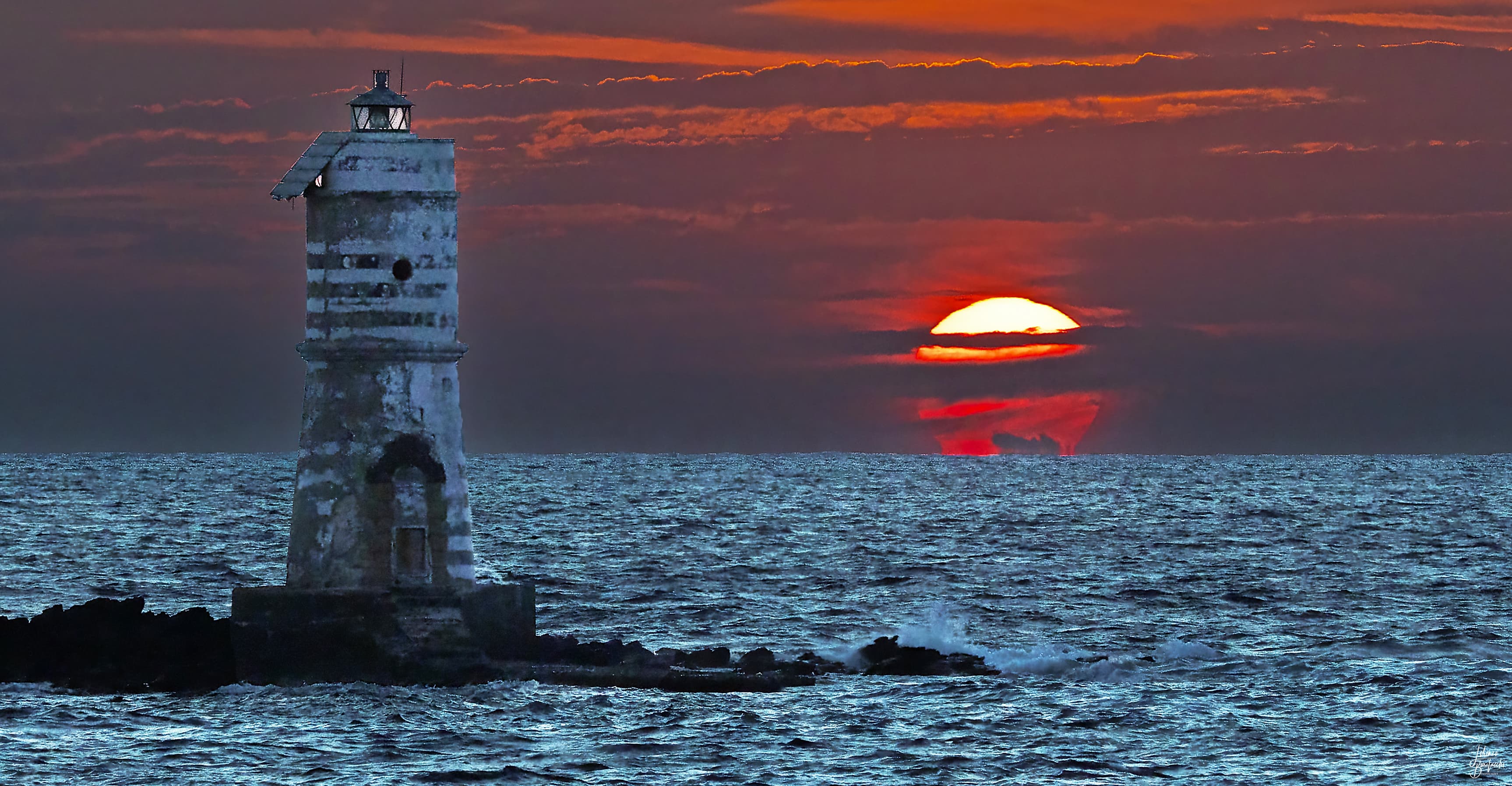 منارةٌ عتيقة في المقدّمة فوق صخور تتوسّط بحراً وخلفها سماء مغيب تتخلّلها غيومٌ وتظهر فيها شمسٌ محمرّة آفلة