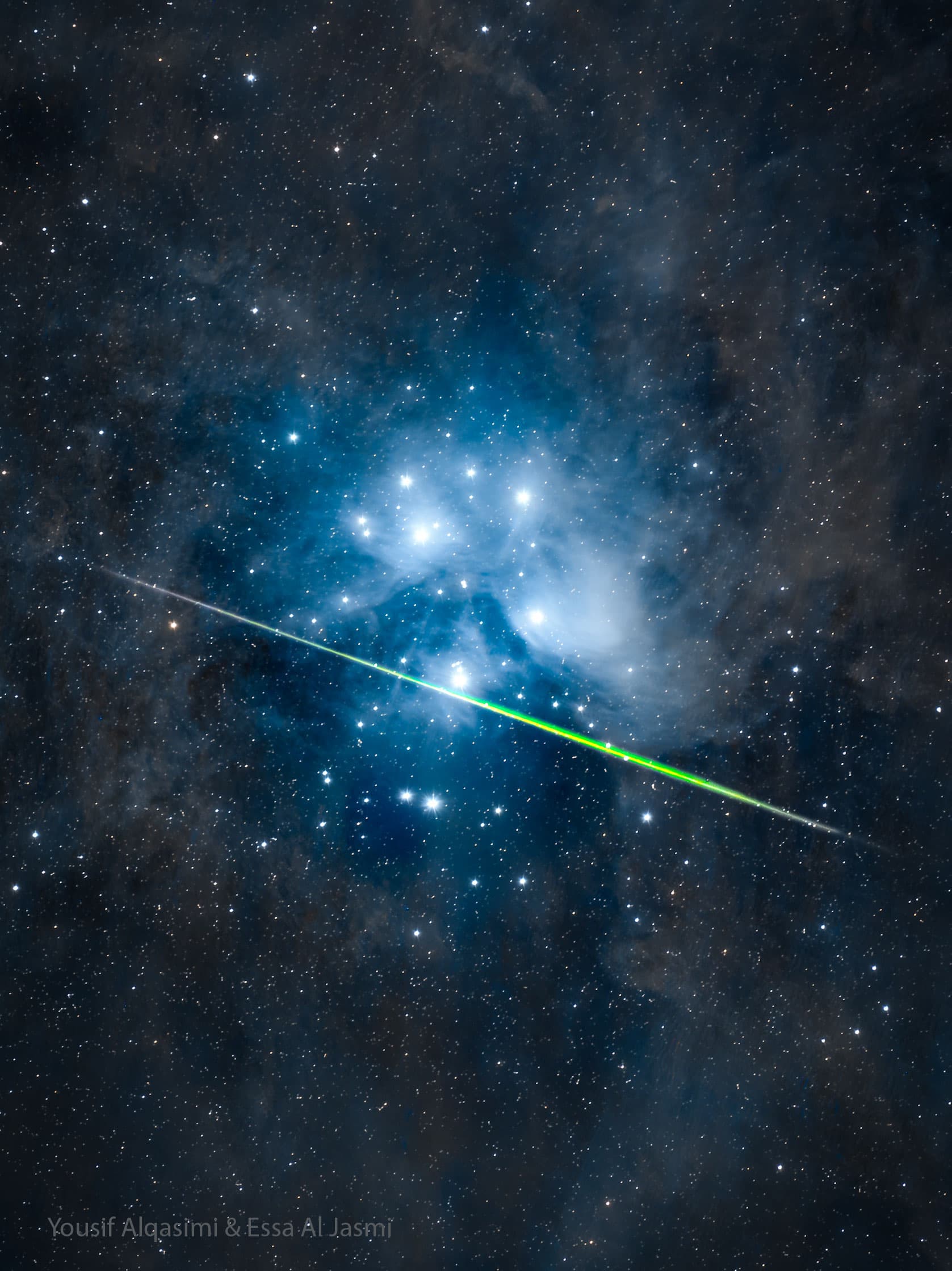 حقلٌ نجميٌّ يحيط بالنجوم الزرقاء الساطعة لعنقود نجوم: عنقود الثريّا النجميّ. يمرُّ عبر العنقود بشكلٍ أفقيٍّ تقريباً خطٌّ أخضرٌ ساطع هو على الأرجح شهاب.