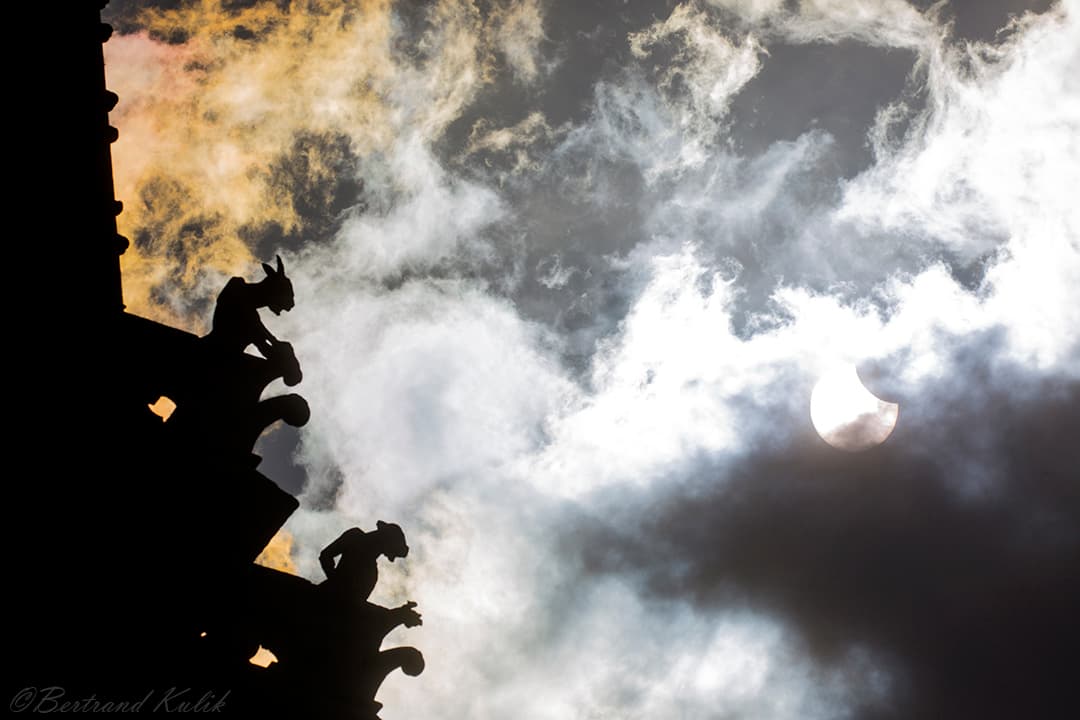 سماء نهارٍ غائمة، وتظهر خلف الغيوم يميناً شمسٌ مكسوفة جزئيّاً بالقمر، بينما يظهر يساراً جانب بناءٍ على سطحه تماثيل گرگول تظهر ظليلة أمام السماء المنيرة.