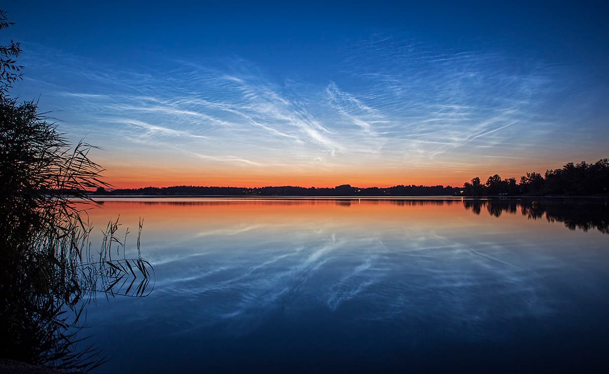 أفق قريب بتضاريس بسيطة يعلوه شفقٌ برتقالي تتفرّع منه سحب رقيقة بيضاء أشبه بعروق عبر سماءٍ زرقاء. ينعكس المشهد على مسطّح مائي في المقدّمة.