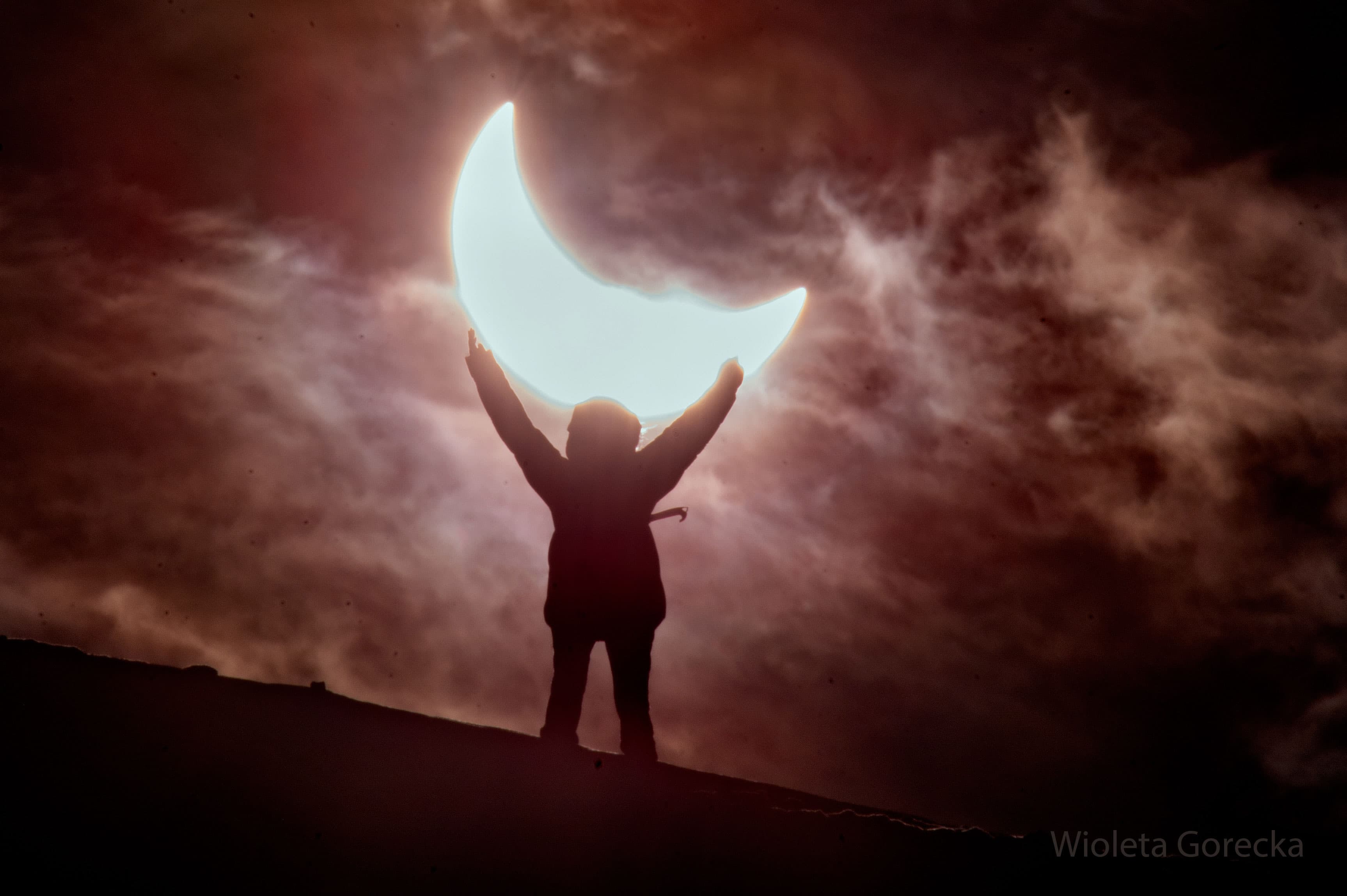 سماءٌ غائمة تظهر بتدرُّج لونٍ ورديٍّ-أحمر بالمجمل. تبدو الشمس مكسوفة جزئيّاً فوق تلّة مائلة. يقف شخصٌ على التلّة ويرفع ذراعَيه للأعلى ويبدو وكأنّه يحمل الشمس المكسوفة جزئيّاً.