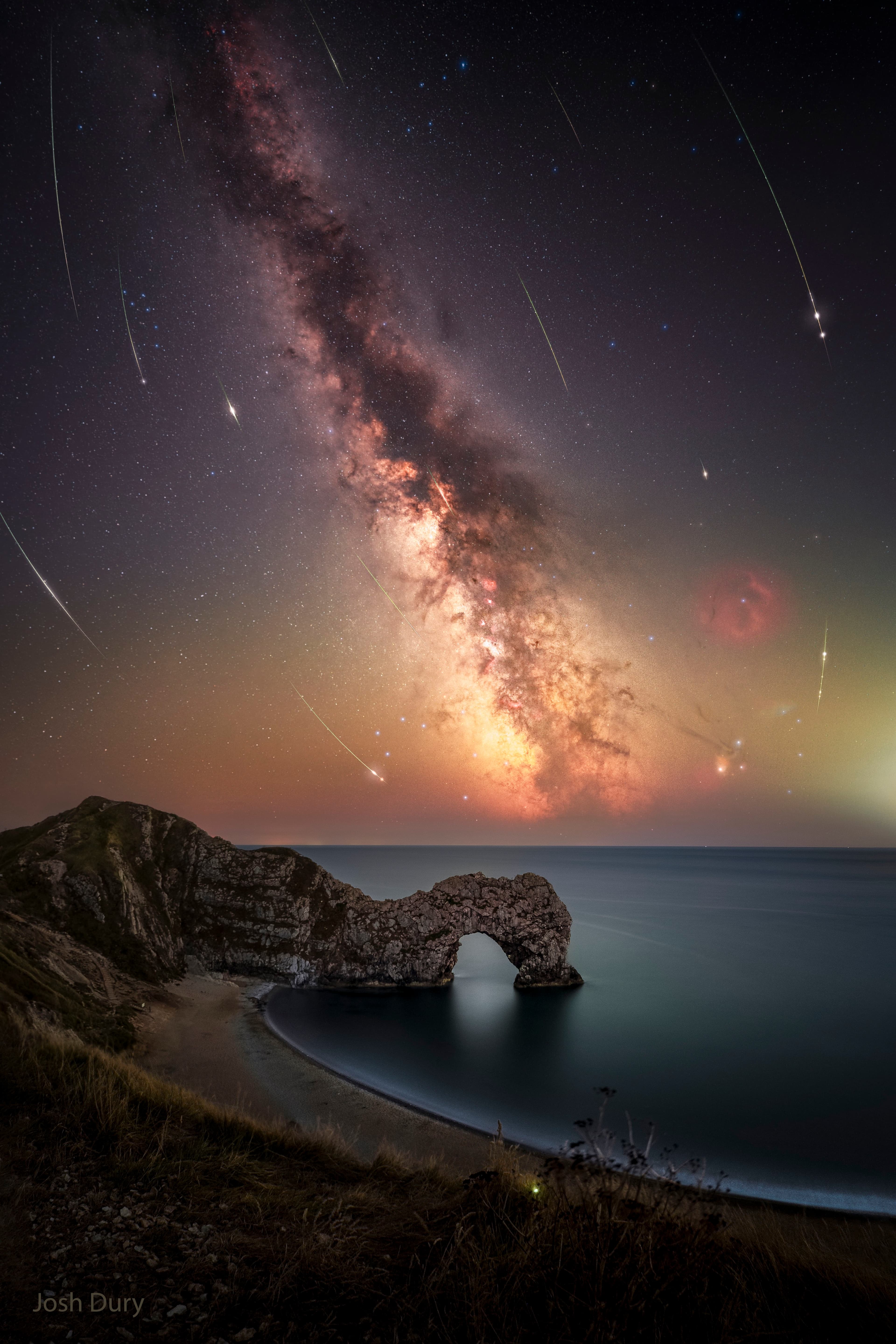 سماءٌ عميقة مع نطاق مجرّتنا درب التبّانة ممتدّاً من أعلى اليسار إلى أسفل اليمين. تُرى خطوط العديد من الشُهب المُنحنية. في المُقدّمة، يُرى شاطئٌ مع نتوءٍ صخريٍّ غير اعتيادي له فتحة.