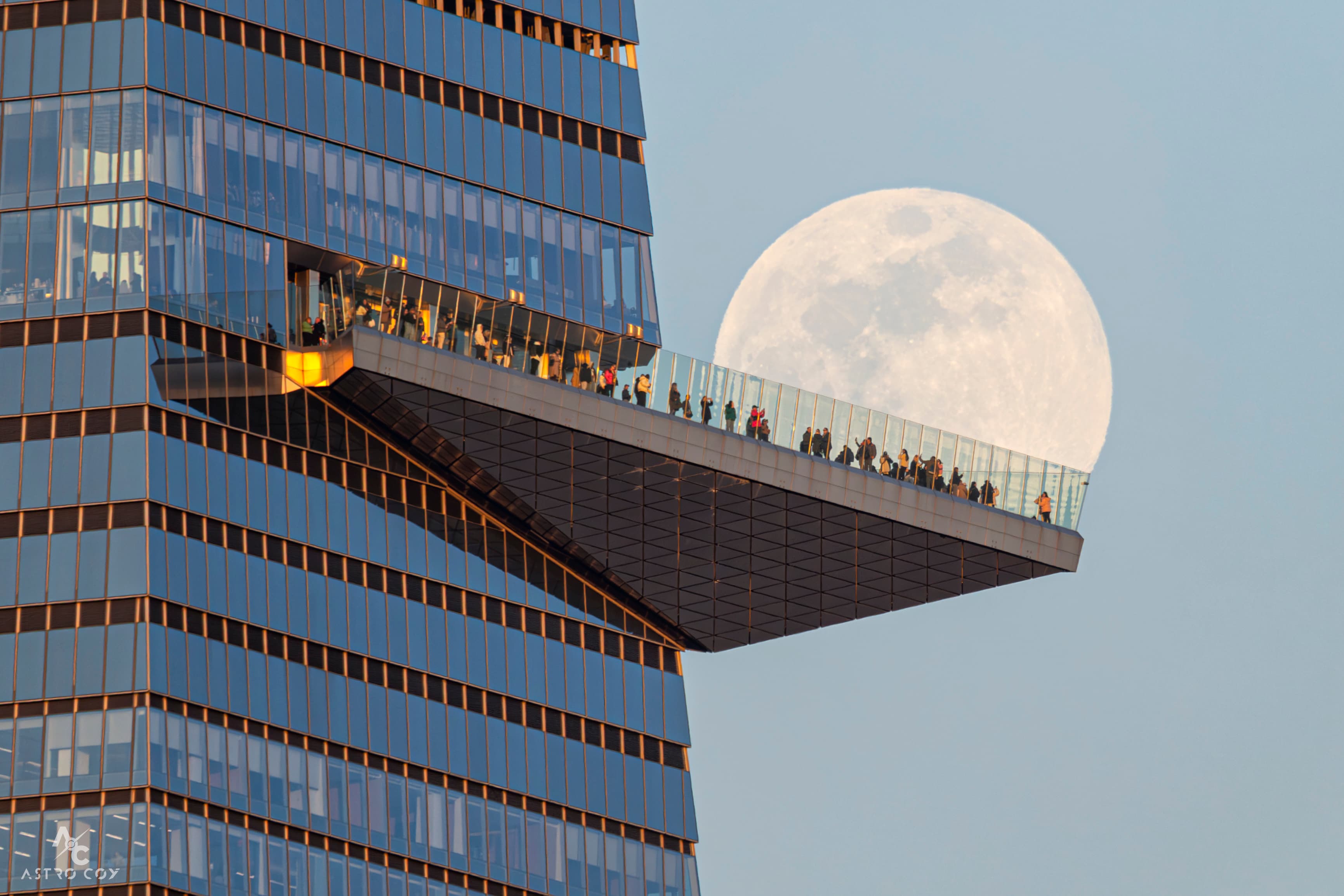 مبنى بواجهات زجاجيّة وأرضيّة ناتئة مسوّرة عليها أناس يتحاذى وراءها جزءٌ مكتملٌ من قمرٌ ساطع يظهر كبيراً لأن الصورة مقرّبة