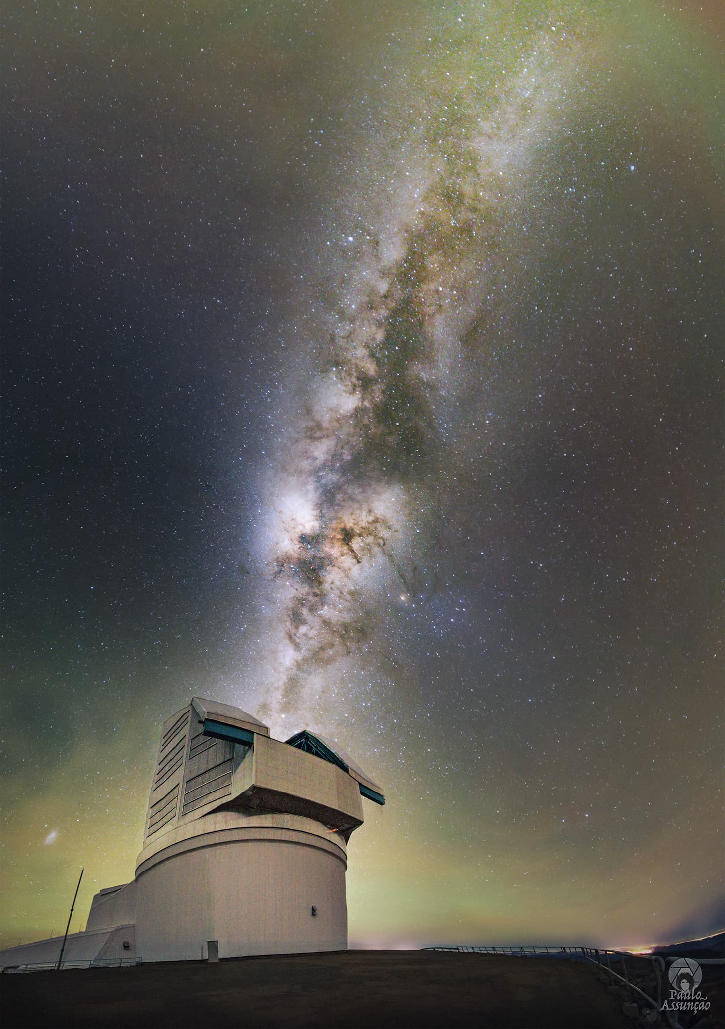 تلسكوبٌ كبيرٌ على اليسار يمتدُّ منه شريطُ مجرّتِنا دربِ التبّانةِ إلى أعلى اليمينِ فيما يُظهِرُ الأفقُ توهّجاً خفيفاً.