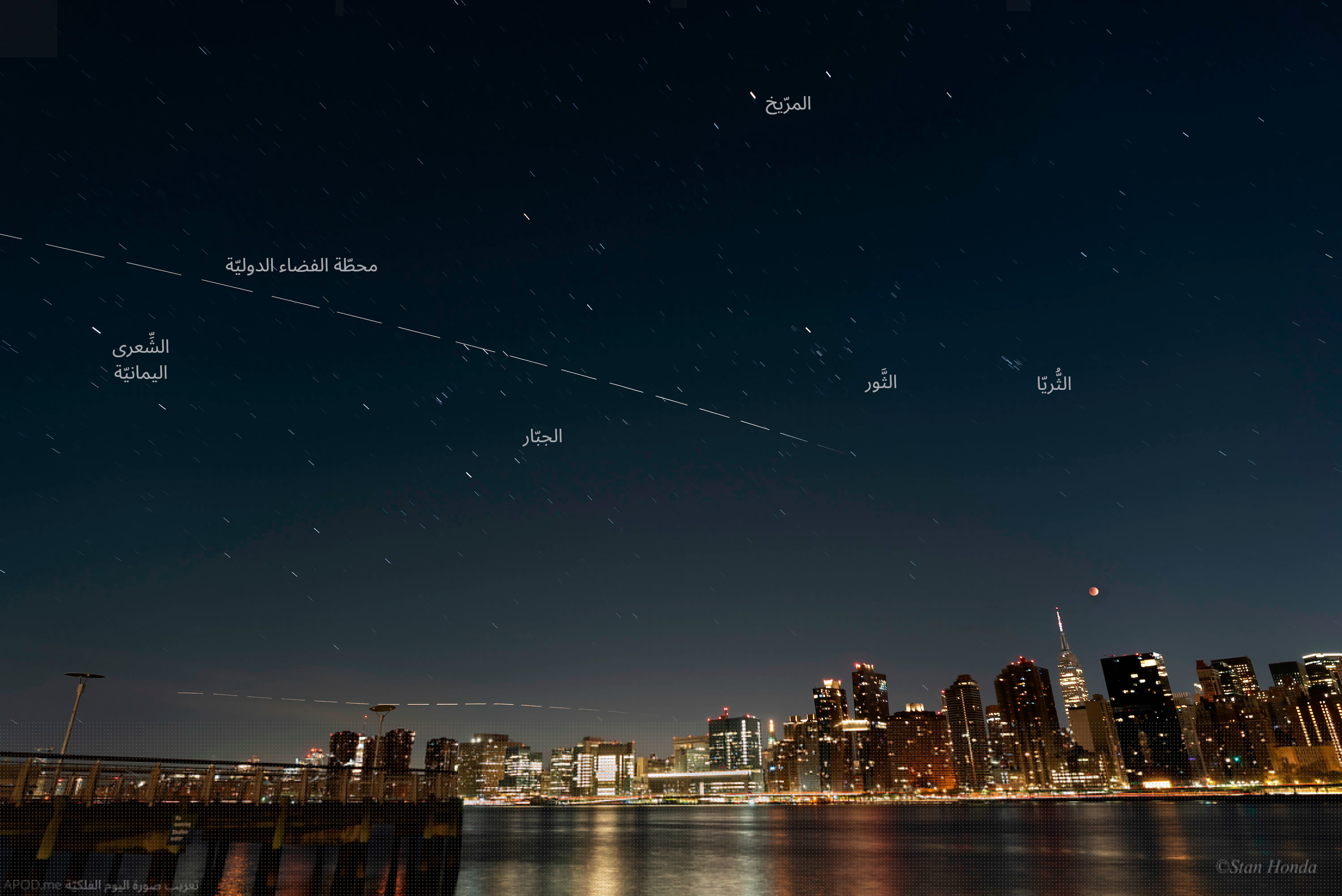 مدينة بخطّ أفق أبنيتها المنارة وفوقها قمرٌ مخسوف وخط متقطّع هو محطّة الفضاء الدولية مع عنونة المرّيخ والثريّا والشعرى اليمانيّة وكوكبتي الجبّار والثور 