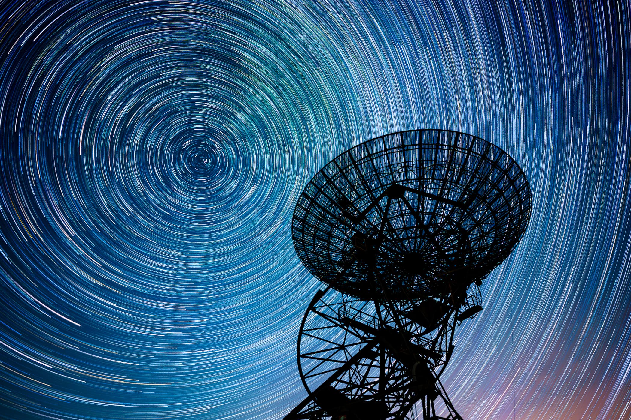 طبق تلسكوب يظهر كشبكةٍ ظليلة على دعامات ومن وراءه السماء تتقوّس فيها مسارات النجوم متّحدة المركز